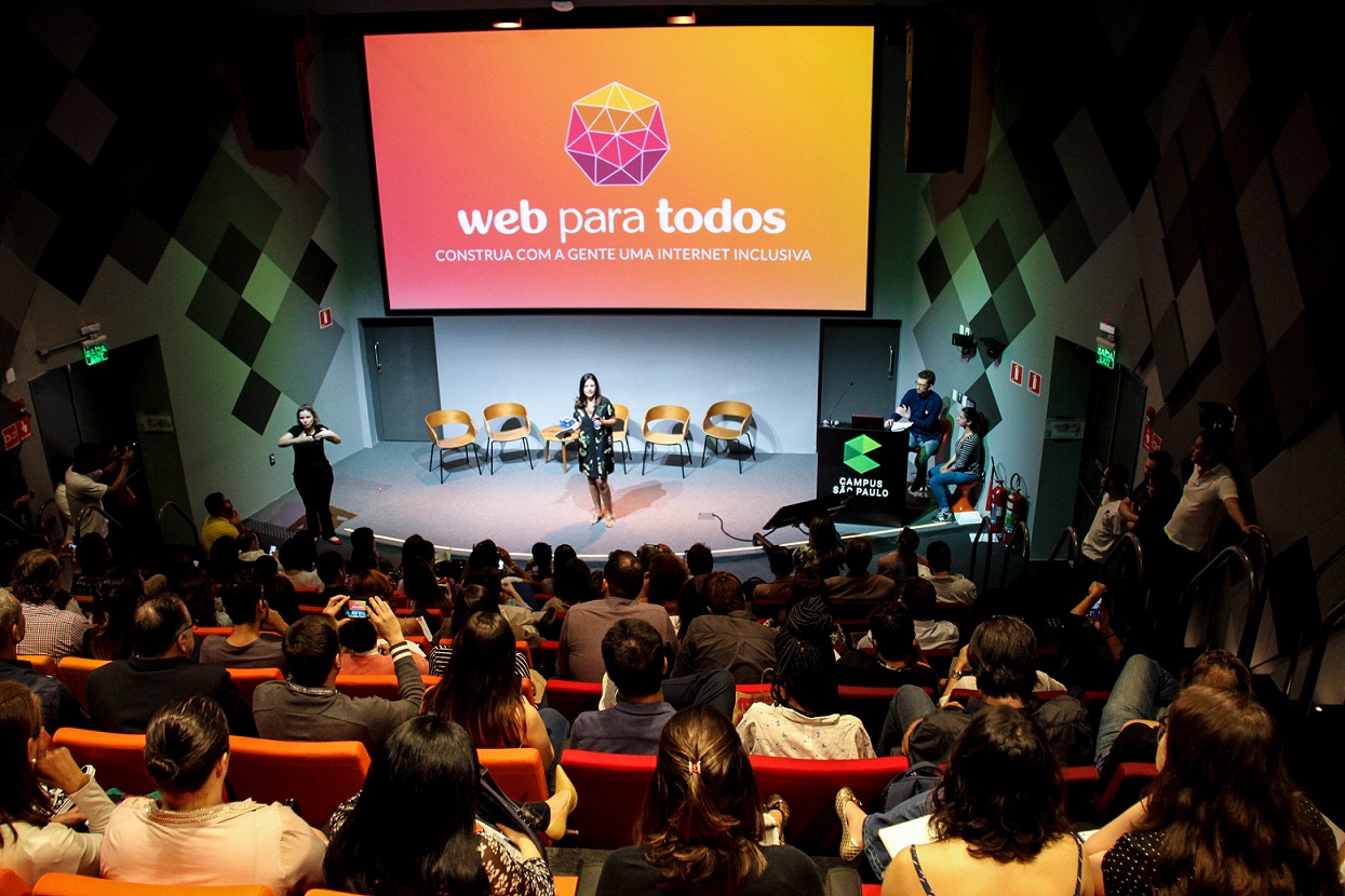 Foto de um auditório lotado, visto de cima. No palco, há uma mulher morena que fala ao público, com uma tela ao fundo que projeta o logo do Movimento Web para Todos.
