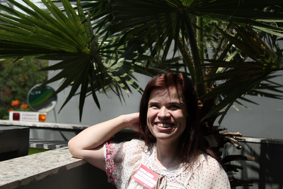 Foto de uma mulher branca com cabelos longos sorrindo. Ela está com o braço direito apoiado em um muro baixo. A mão direita apoia a cabeça dela.