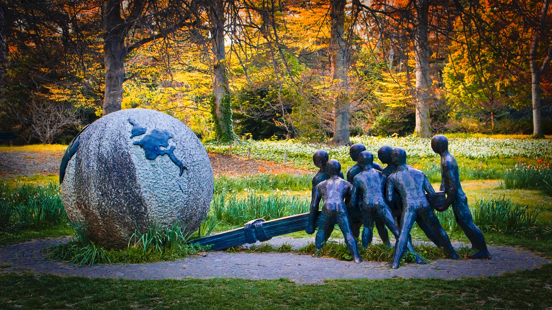 Foto de um ambiente arborizado e gramado, onde há uma escultura. A escultura representa um globo terrestre e sete pessoas somando forças para movê-lo utilizando uma alavanca.