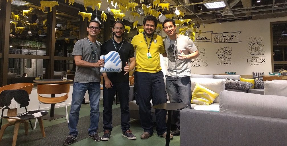 Foto de quatro homens em pé sorrindo para a câmera em um ambiente fechado, amplo, com sofás e cadeiras. Um deles segura uma almofada com o símbolo da ProDeaf.