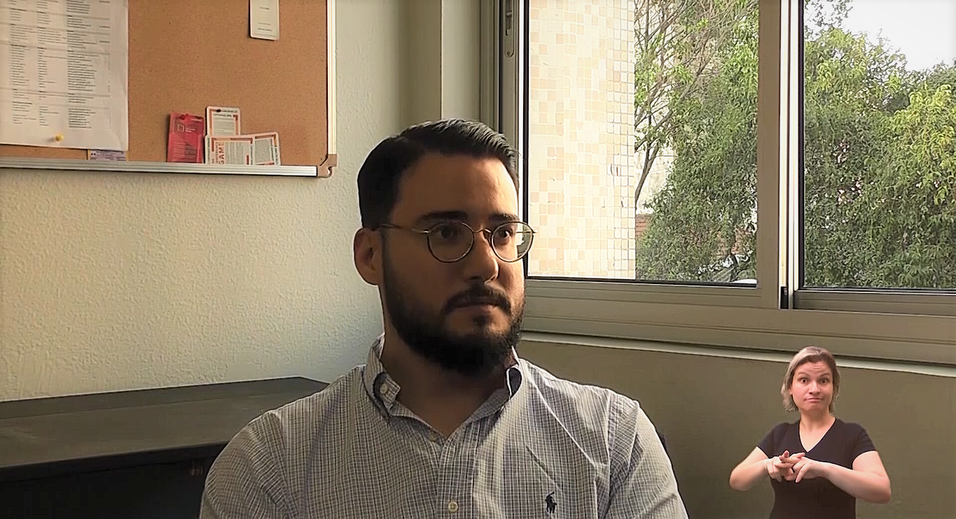 Foto de um homem jovem, moreno, com barba e óculo. Ele usa uma blusa social.
