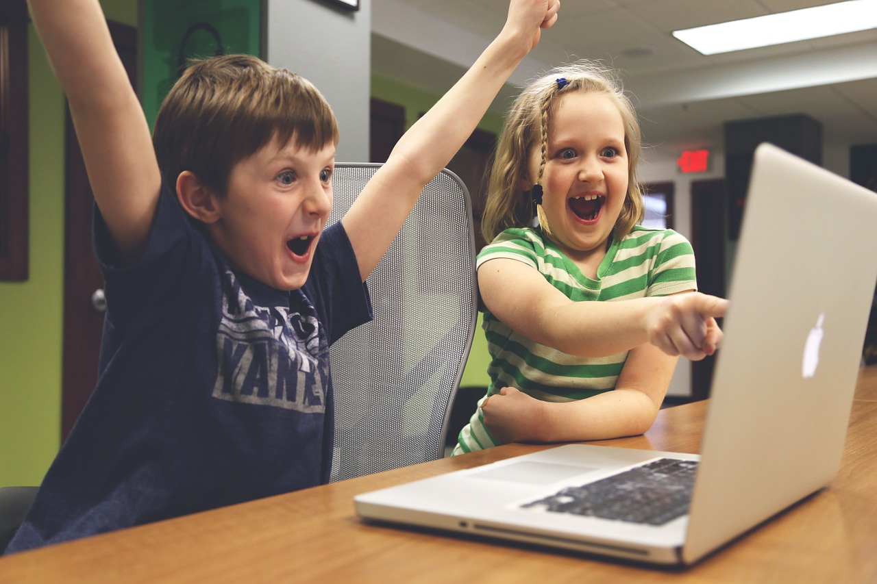 Foto de uma menina e um menino sentados na frente de um laptop. Eles olham para a tela com expressões faciais que demonstram muita alegria.