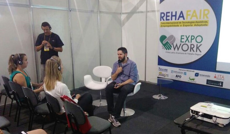 Foto do William sentado com um microfone na mão na frente de duas mulheres sentadas. Ao fundo, há um banner com o texto REHAFAIR.