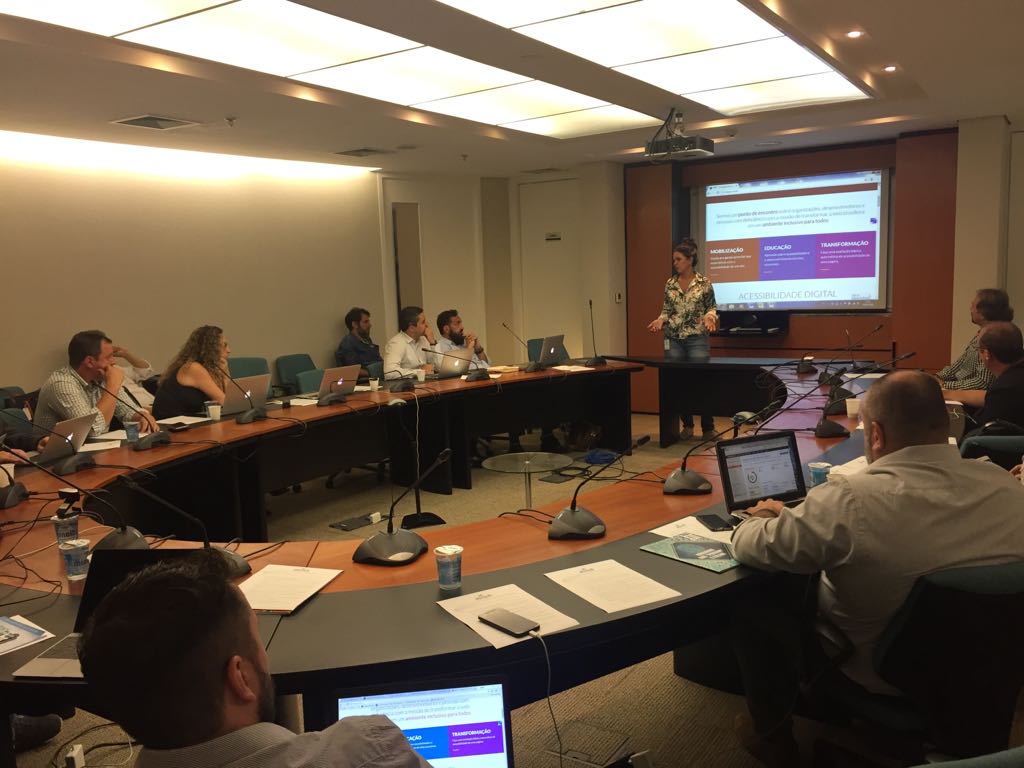 Foto de uma sala com pessoas sentadas observando Simone em pé. Ao fundo, há um telão com a página principal do Web para Todos.