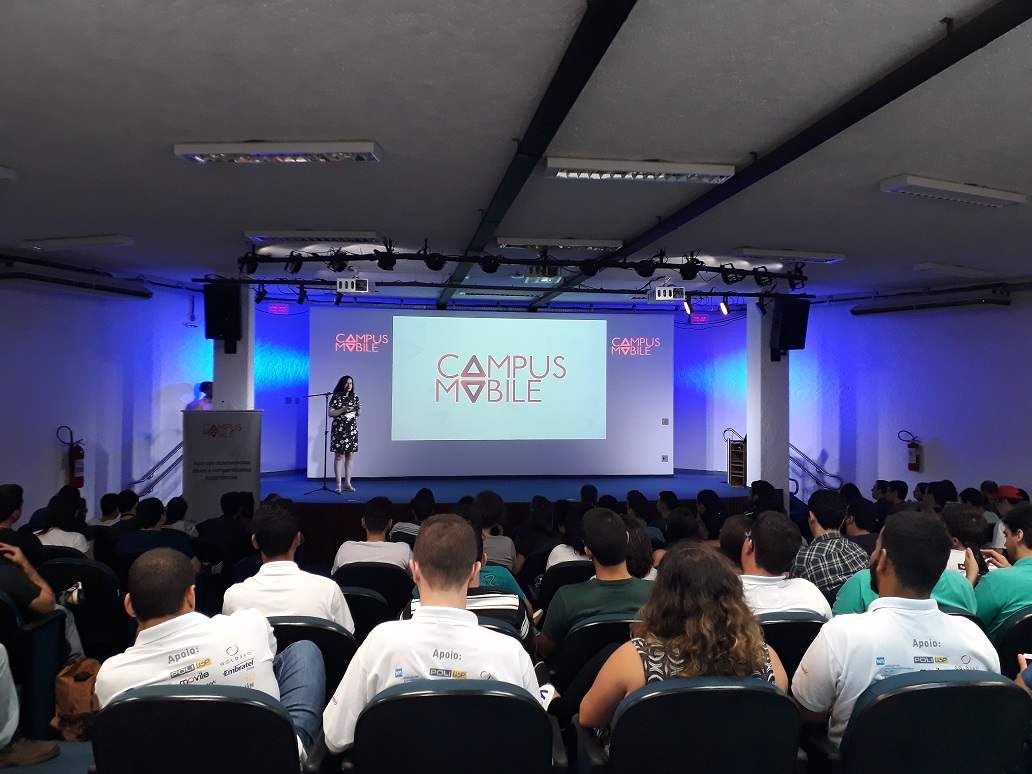 Foto de uma mulher em pé em um palco na frente de uma platéia lotada. Ao fundo, atrás da mulher, há um telão com o logo da Campus Mobile.