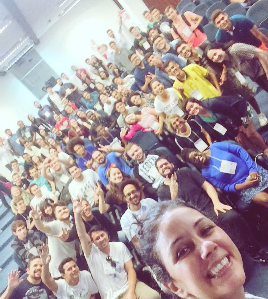 Foto estilo selfie da Simone com participantes do Campus Mobile ao fundo sorrindo. Alguns estão com braços levantados fazendo sinal de V e coração com as mãos.