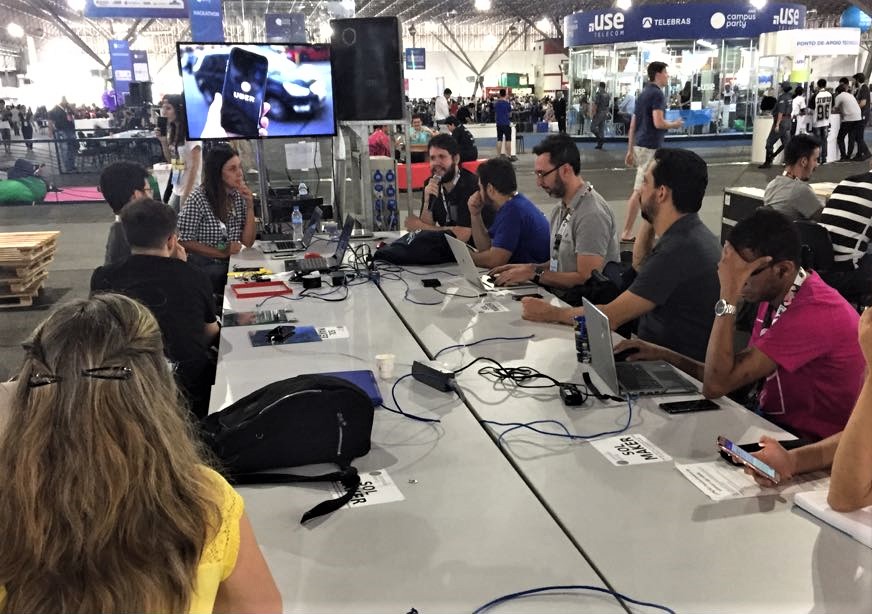 Foto de um espaço aberto com pessoas reunidas ao redor de uma mesa retangular com laptops. Na ponta da mesa, William seguram microfones e falam para os participantes.
