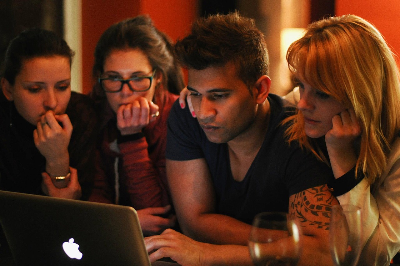 Foto de quatro pessoas olhando a tela de um laptop. Há três mulheres e um homem com uma das mãos no teclado.
