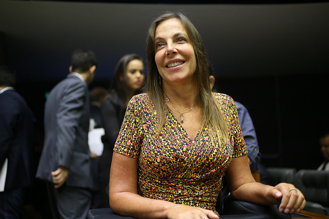 Foto da Mara Gabrilli. Ela é branca, com cabelos longos e claros e está sorrindo, sentada.