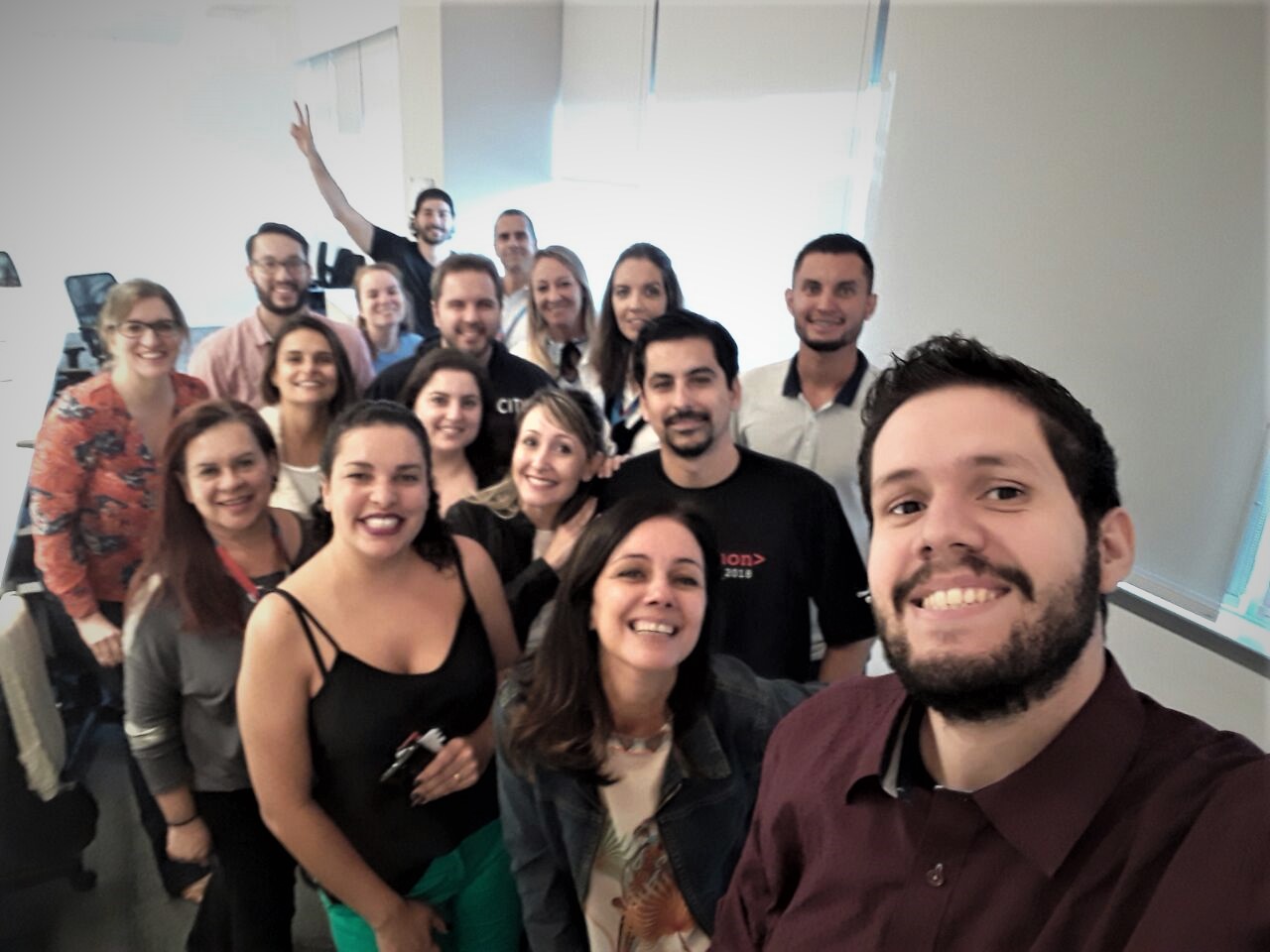 Homens e mulheres estão em pé sorrindo ao lado de uma mesa branca. Eles olham para a mesma direção e estão em uma sala fechada.