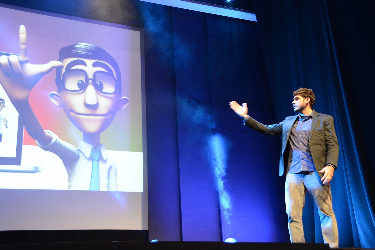 Foto de um homem branco de cabelos curtos que veste um paletó e camisa na cor cinza e calça jeans em cima de um palco apontando com a mão direita aberta para um telão. Nele, há o intérprete virtual da Hand Talk.
