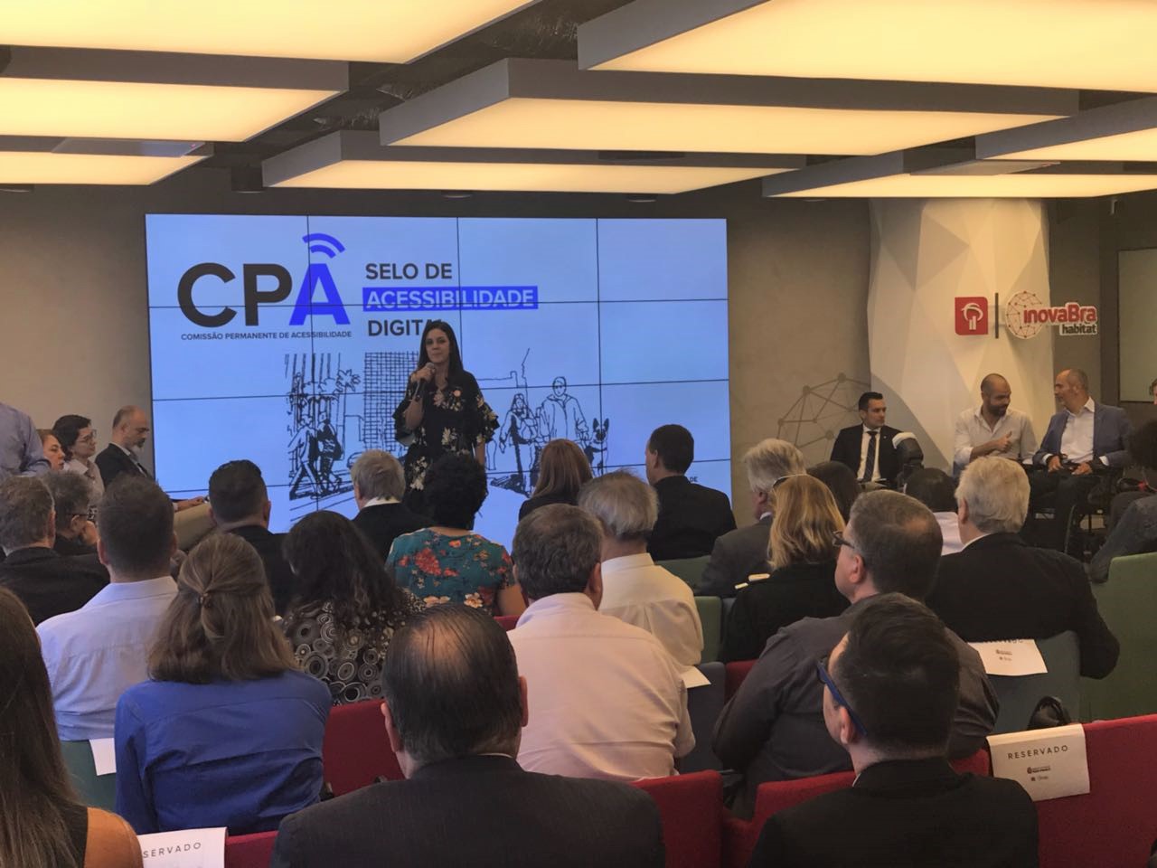 Foto da Simone Freire em pé, segurando um microfone na frente de um público sentado em uma sala fechada. Ao fundo, há um telão com o texto "CPA - Comissão Permanente de Acessibilidade. Selo de Acessibilidade Digital".