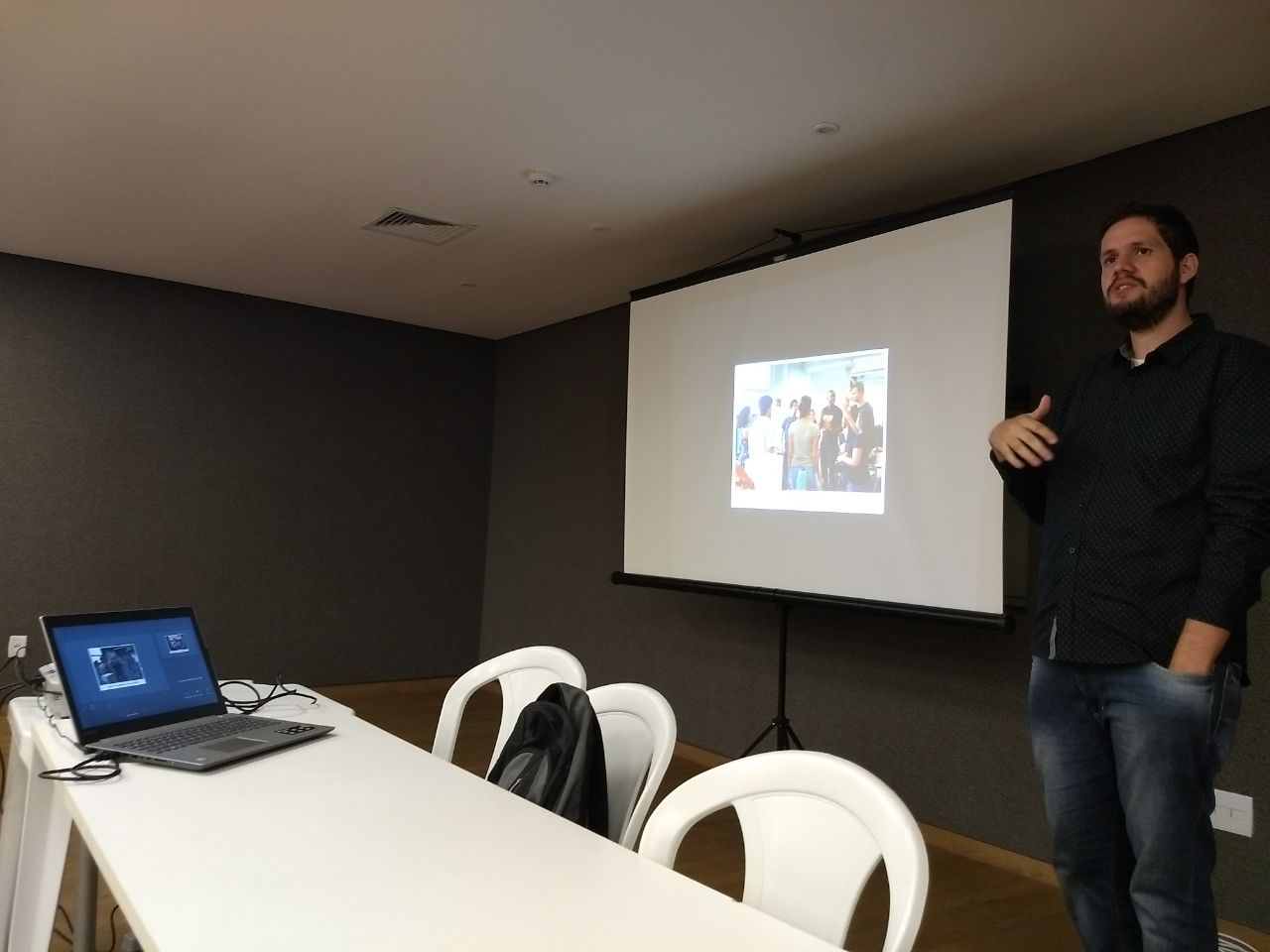 Foto do William em pé gesticulando e com uma das mãos no bolso. Ao fundo, há um telão. 