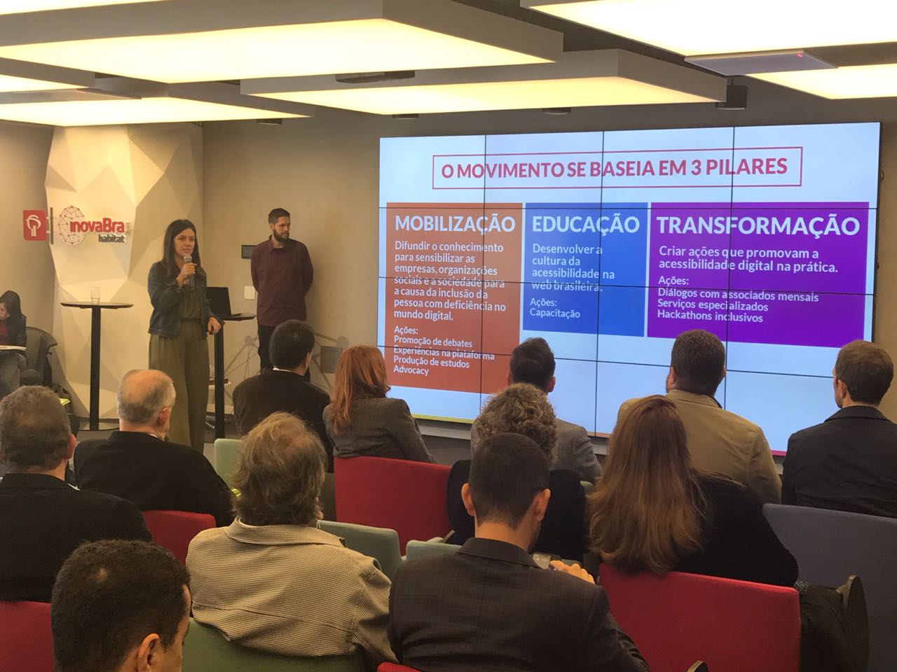 Foto de uma mulher e um homem em pé na frente de um público. Ela está segurando um microfone e o homem está olhando para o slide. Eles estão em uma sala fechada. Ao fundo, há um telão com um texto em destaque: "O movimento se baseia em três pilares: mobilização, educação e transformação".