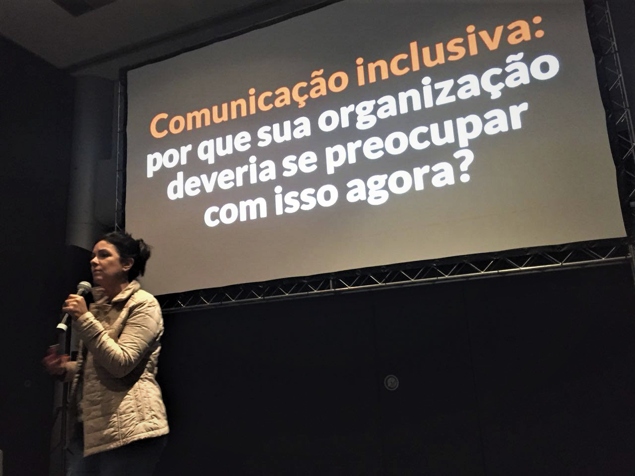 Foto do auditório fechado. Ao fundo, o telão com o nome da palestra ao fundo e uma mulher ao microfone. Ela é alta, branca, tem cabelos escuros presos em um coque e olha para o lado esquerdo.