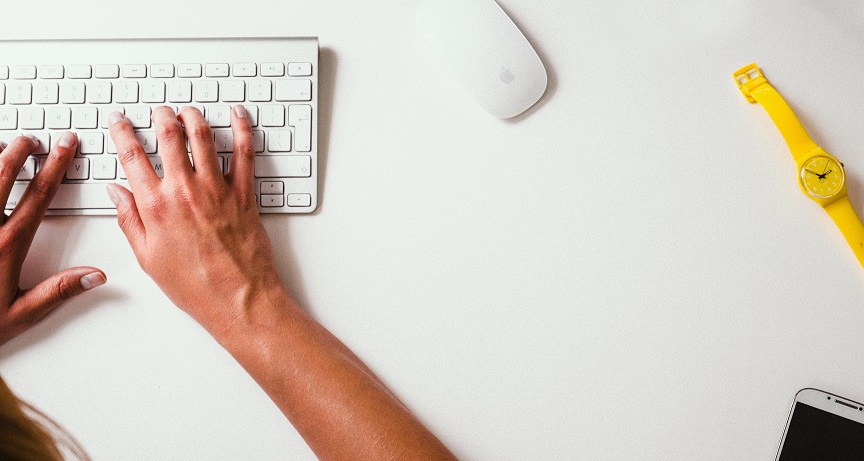Foto de duas mãos em um teclado branco. Há um mouse branco e um relógio amarelo ao lado.
