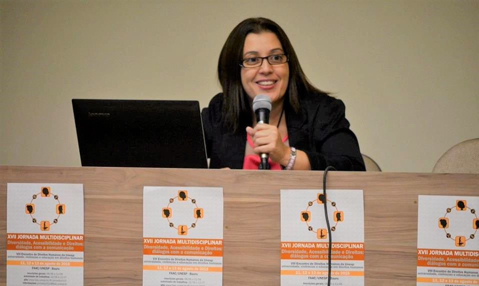 Lucinéa sentada falando ao microfone. Ela tem cabelos longos e lisos e está usando óculos.