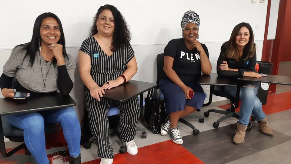 Rubi de la Fuente, Flavia Durante, Michelle Fernandes e Simone Freire sorrindo. Elas estão sentadas em cadeiras do tipo estudante em uma sala.