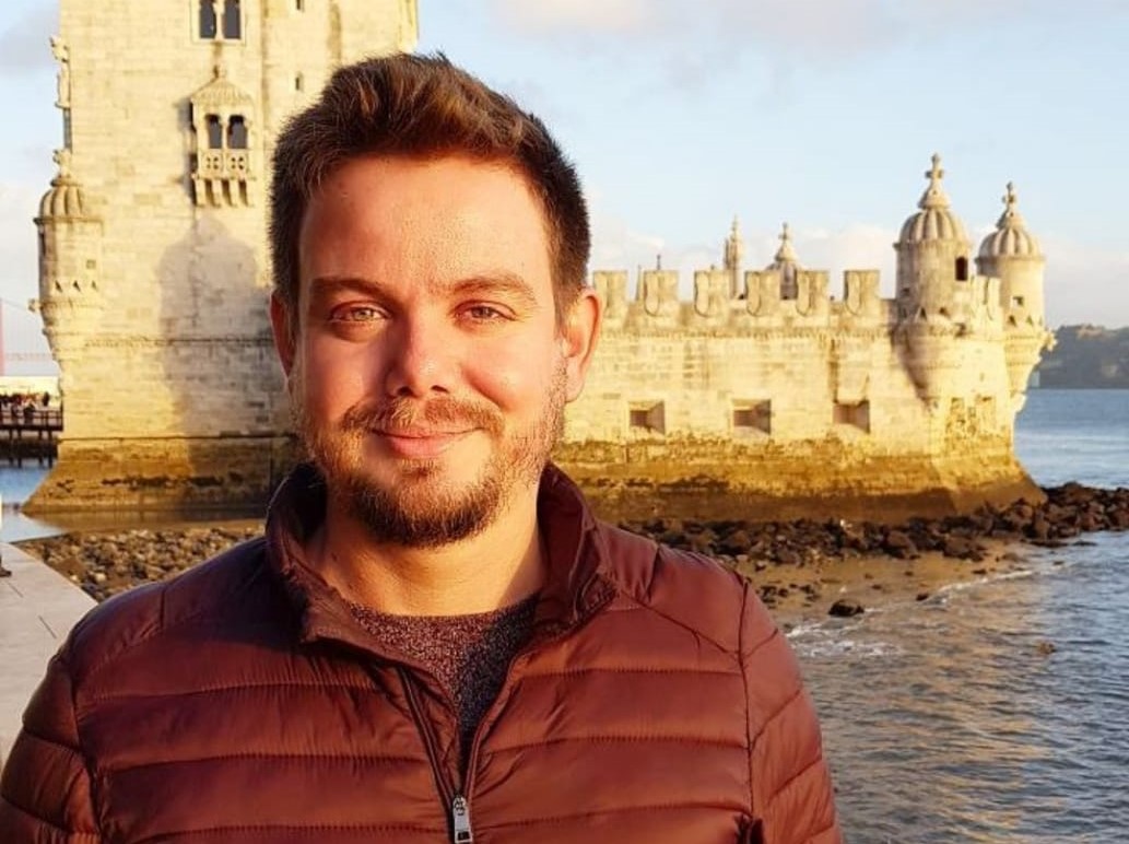 Foto do Valter Lenine Fernandes sorrindo. Ele está em um ambiente externo, próximo ao mar. Ao fundo, há um castelo. Ele é loiro, tem olhos claros e cabelos curtos.