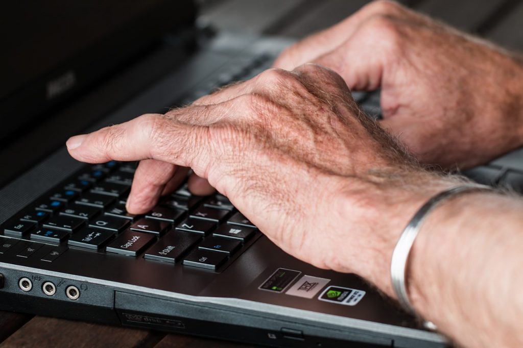 Mãos de uma pessoa idosa digitando no teclado de um computador. 