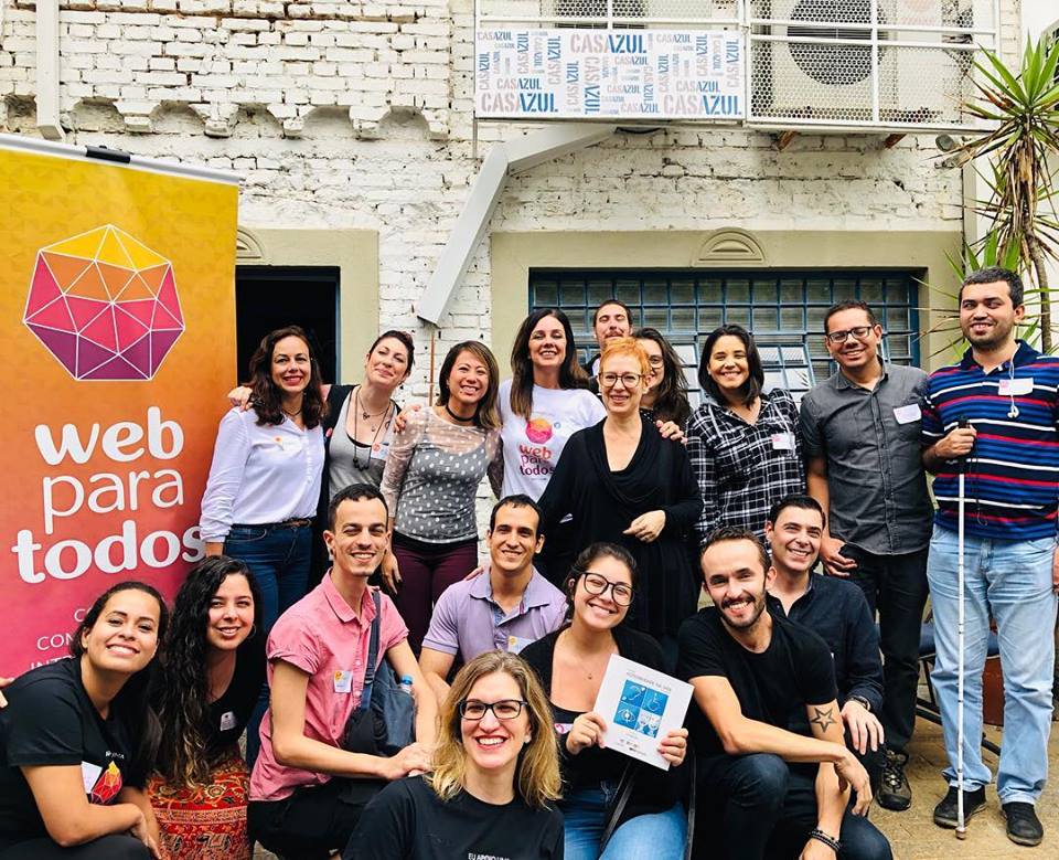 Grupo com 18 pessoas sorrindo na área externa de uma casa. Algumas pessoas estão em pé e outras estão agachadas.