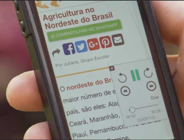 Mão de uma pessoa segurando um celular. A tela mostra uma notícia com a plataforma Audima.