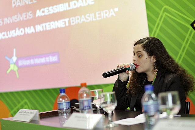 Sarah Marques está sentada, falando ao microfone. Há uma mesa na frente dela com várias garrafas de água. Ela tem cabelos castanhos longos e encaracolados.