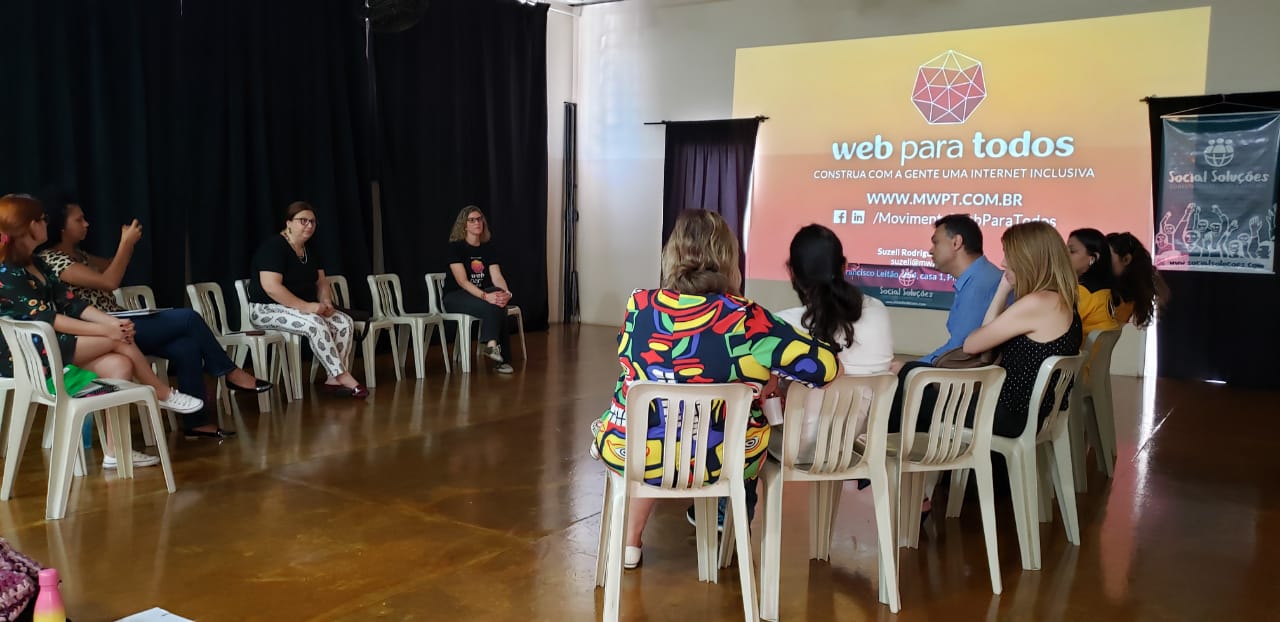 Pessoas sentadas em cadeiras organizadas em roda. Ao fundo, há uma imagem grande projetada na parede com o logo do Movimento Web para Todos e dois banners do Social Soluções em cada lado.
