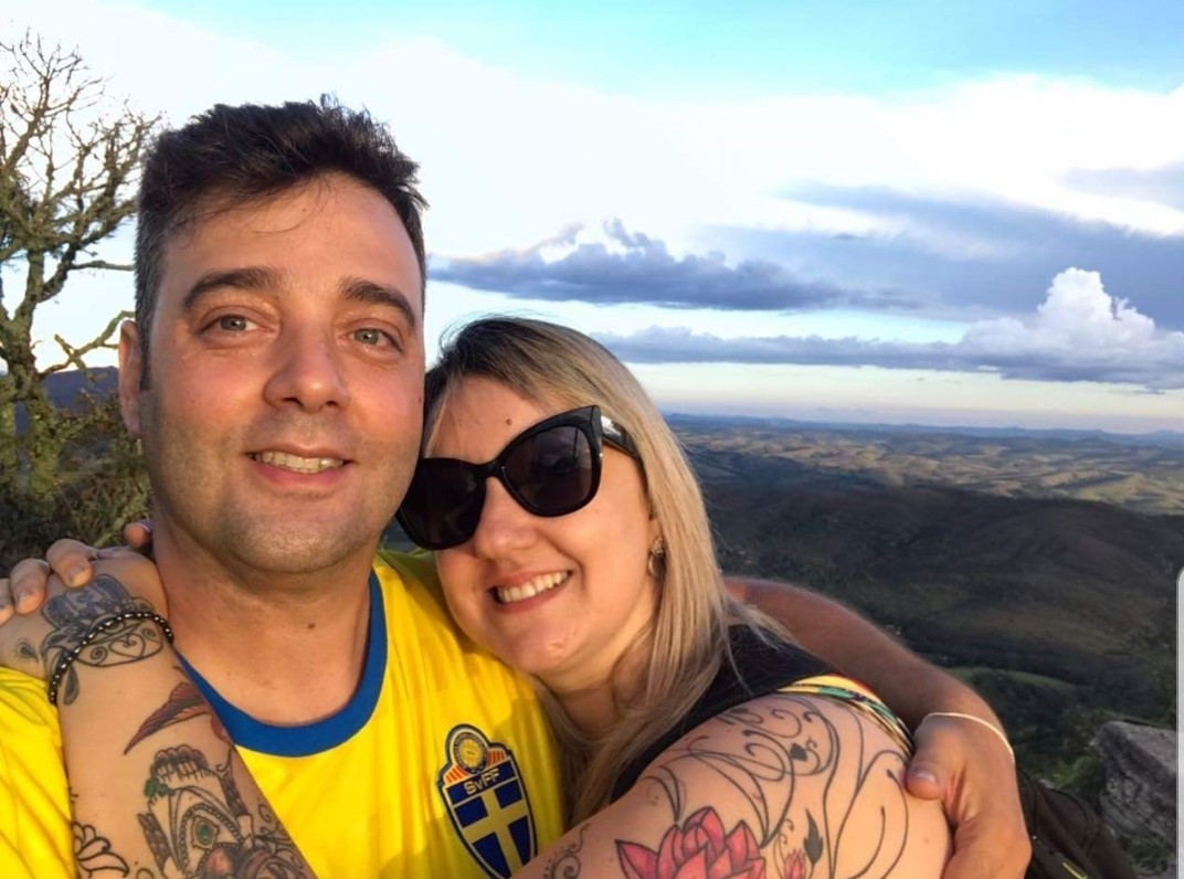 Matheus e Karina estão abraçados em uma área externa em um dia ensolarado. Ambos sorriem. Ao fundo, há uma área verde.
