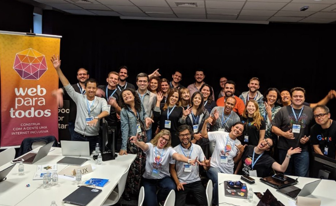 Grupo com aproximadamente 30 pessoas reunidas sorrindo, fazendo gestos com as mãos.