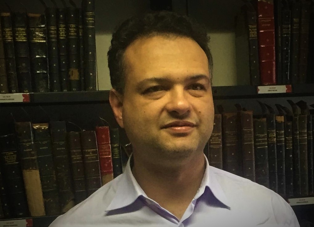 Carlos Ferrari com leve sorriso no rosto. Ele tem cabelos curtos castanhos escuros. Ao fundo, há uma estante com livros.