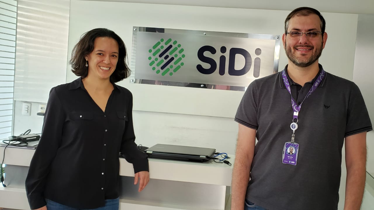 Fernanda Kussama Pellegrini e Marcelo Anjos estão em pé, sorrindo, em um ambiente fechado. Ao fundo, há uma placa com o nome "SiDi".