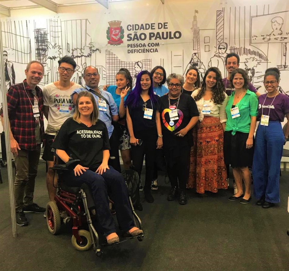 Grupo com mais de 10 pessoas com e sem deficiência, em pé, sorrindo. Elza Maria faz parte desse grupo. Ela tem cabelos longos encaracolados e está usando uma saia longa vermelha com uma blusa branca. Ao fundo, há um banner com ilustração de prédios e pessoas com o texto: "Cidade de São Paulo - Pessoa com Deficiência".