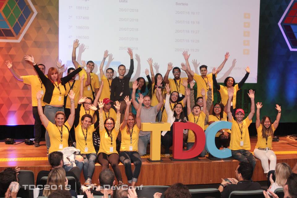 Grupo de pessoas em pé e sentadas em cima de um palco ao redor das letras TDC. A maioria está vestida com uma blusa amarela e está sorrindo com as mãos para cima.