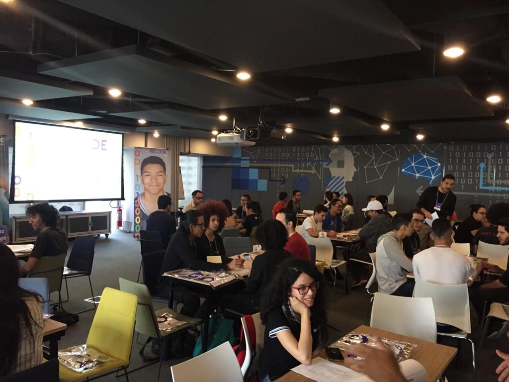 Grupos de pessoas sentadas ao redor de diversas mesas em uma sala fechada. Ao fundo há um telão e um banner com um homem sorrindo.