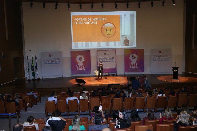 Simone Freire está em pé, falando ao microfone em um palco na frente de diversas pessoas que estão sentadas.