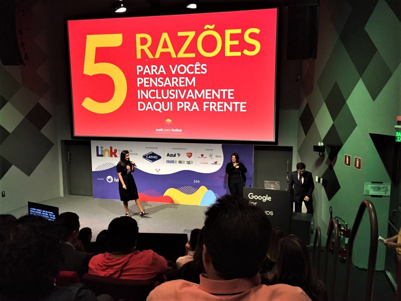 Simone Freire está em pé, segurando um microfone no palco do auditório do Google Campus São Paulo.