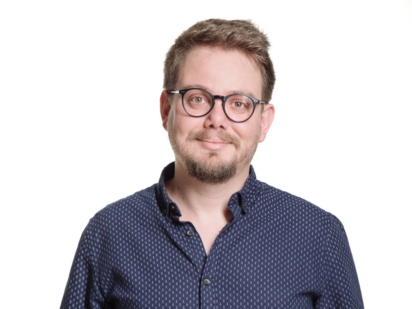 Valter Lenine sorri. Ele é loiro, tem cabelos curtos e barba. Está usando óculos e uma blusa social estampada e azul.