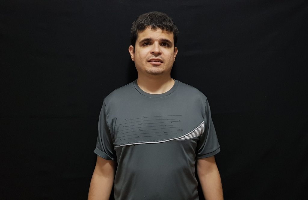 Foto do Diniz Candido em pé com leve sorriso. Ele tem cabelos curtos, castanhos e está usando uma blusa na cor cinza.