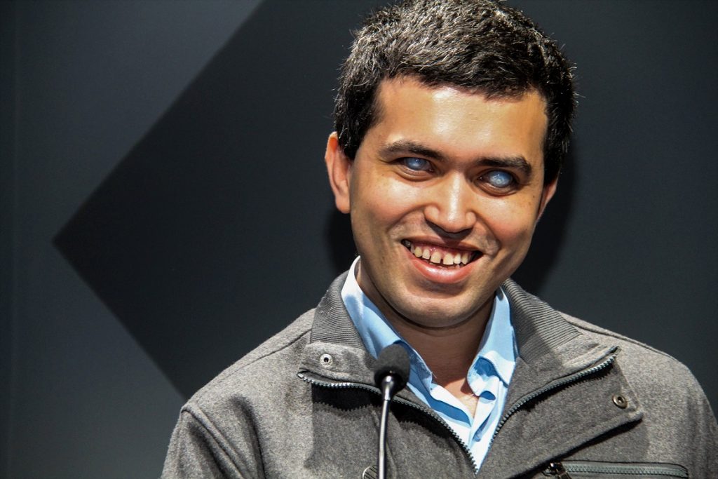 Leonardo sorri. Ele tem olhos esbranquiçados, cabelos curtos e negros e usa casaco cinza e blusa azul.