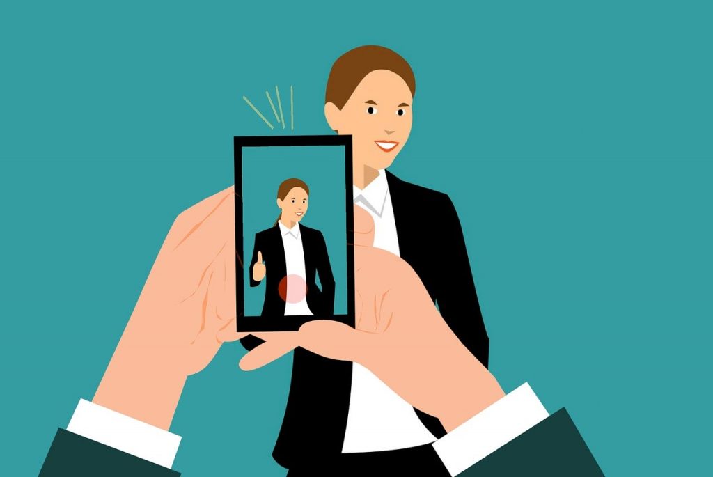 Ilustração das mãos de uma pessoa segurando um smartphone na direção de uma outra que está em pé, olhando na direção do equipamento. A tela dele mostra a imagem da pessoa que sorri e faz o sinal de positivo com uma das mãos. Ainda na tela, há um círculo vermelho no rodapé.