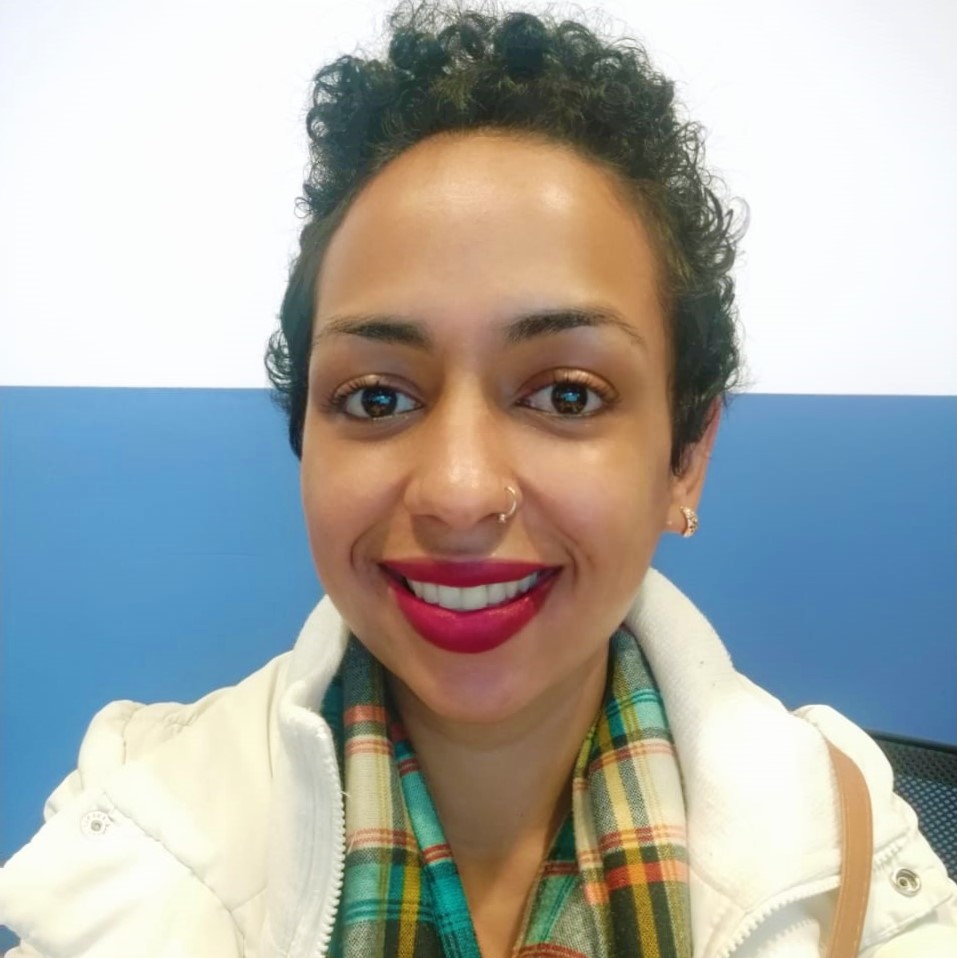 Foto da Nayara sorrindo. Ela tem cabelos encaracolados castanhos e curtos. Usa um brinco pequeno, um piercing no nariz e batom vermelho.