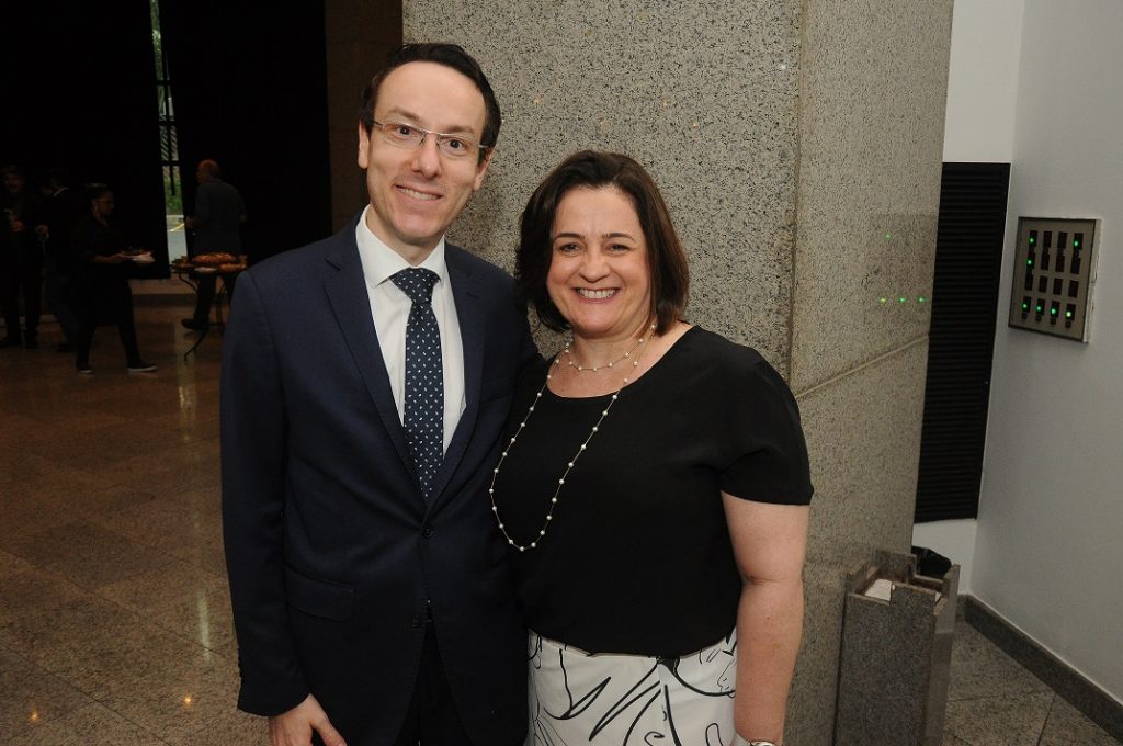 Foto com César Eduardo ao lado da Denise. Ambos sorriem e estão em pé em uma área fechada. Ele tem cabelos curtos, usa óculos e usa terno e gravata. Ela tem cabelos lisos, acima dos ombros e usa blusa preta com colar grande.