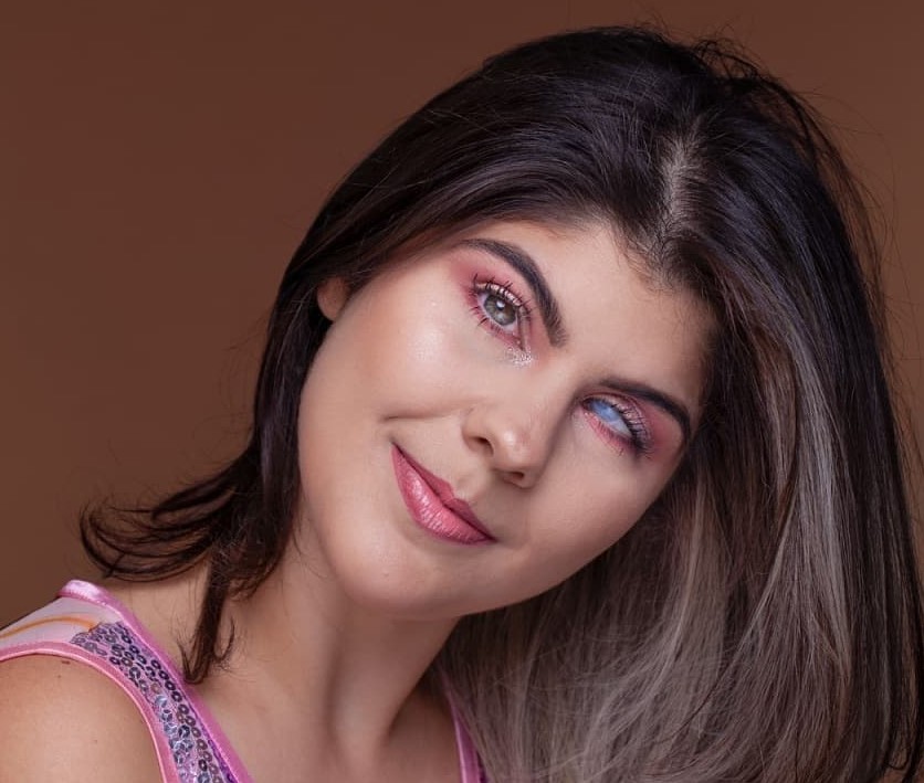 Foto da Isa Meirelles com um leve sorriso. Ela tem cabelos lisos e longos, castanhos claros, na altura dos ombros, tem os olhos claros. Um deles é azul esbranquiçado. Isa usa uma blusa rosa com lantejoulas brilhantes.