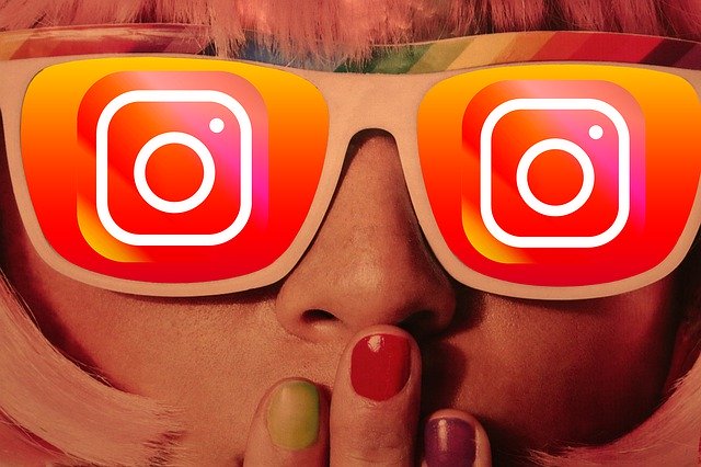 Ilustração de uma pessoa loira com óculos, que estão com logo do Instagram em cada lente na cor laranja em degradê. Ela está com as mãos abaixo do nariz, com unhas pintadas com cores diversas.