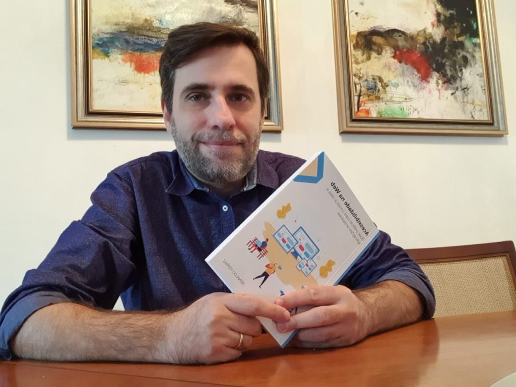 Foto do Reinaldo Ferraz sentado, sorrindo. Ele tem cabelos curtos, barba grisalha e usa blusa social na cor azul. Reinaldo segura com as duas mãos o livro “Acessibilidade na web: boas práticas para construir sites e aplicações acessíveis”. As mãos estão apoiadas em cima de uma mesa. Ao fundo, há dois quadros abstratos na parede.