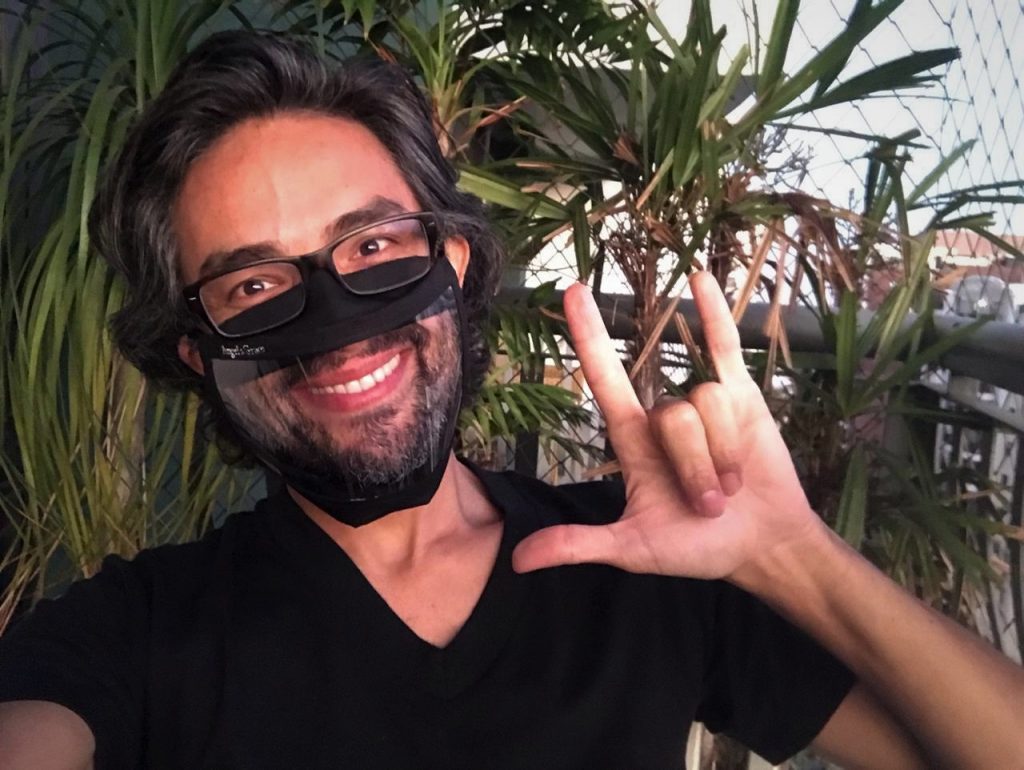 Foto do Alexandre Ohkawa em uma área externa, durante o dia. Ele sorri e faz o sinal "I love you". Ele tem cabelos curtos e lisos, barba grisalha. Usa óculos preto, blusa preta e uma máscara preta com parte transparente na área da boca, que mostra seu sorriso. Ao fundo, há algumas plantas.
