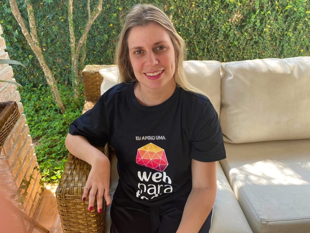 Foto da Nathalia Blagevitch sentada em um sofá, que está em uma área externa, durante o dia. Ela sorri. Nathalia é loira, tem cabelos longos e lisos e usa uma blusa preta com o logotipo do Movimento Web para Todos.