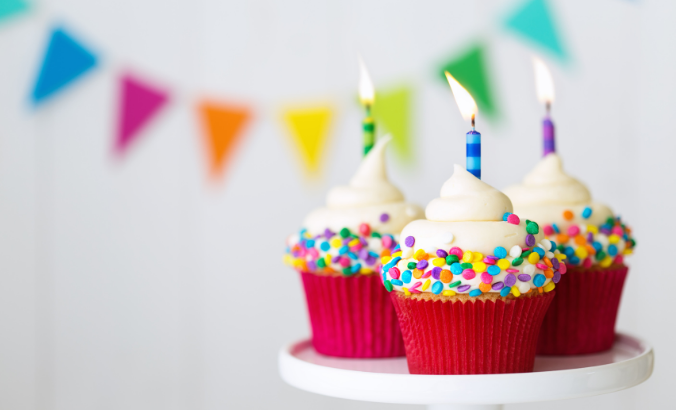 Foto em close de três cupcakes coloridos com velas acesas. Na parede ao fundo, há bandeirinhas de cores variadas desfocadas.