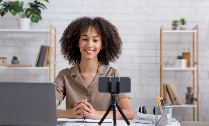 Foto de uma mulher negra sentada na frente de um laptop e de um smartphone que está em um tripé. Ela sorri.